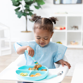 A young curly-haired girl sits in a high chair, wearing a Bumkins Ruffle Apron Bib: Cinderella while eating from a blue plate with utensils.