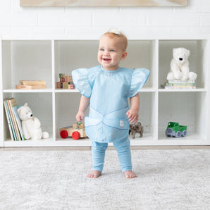 A baby smiles, wearing a Bumkins Ruffle Apron Bib: Cinderella while standing in front of shelves with toys and books.
