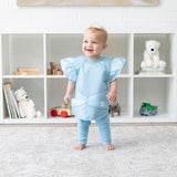 A baby smiles, wearing a Bumkins Ruffle Apron Bib: Cinderella while standing in front of shelves with toys and books.