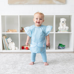 A young girl stands in a bright playroom, wearing a bib from Bumkins Snack In Character 4-Piece Set: Disney Princess Magic Cinderella.