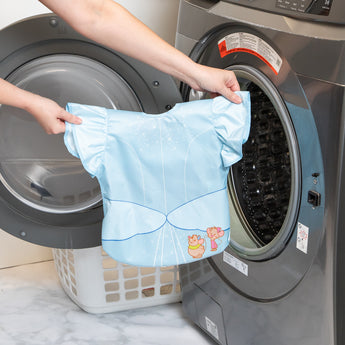A person holds a bib from Bumkins Snack In Character 4-Piece Set: Disney Princess Magic Cinderella in front of an open washing machine.