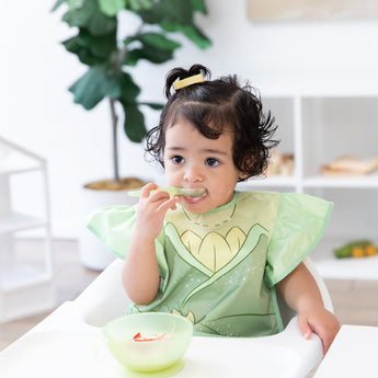 A young girl wears a Bumkins Ruffle Apron Bib: Tiana in a high chair, eating yogurt and berries from a green bowl with a spoon.