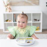 A baby in a Bumkins Ruffle Apron Bib: Tiana sits at a white table with a gray plate of food, looking off to the side with a soft smile.