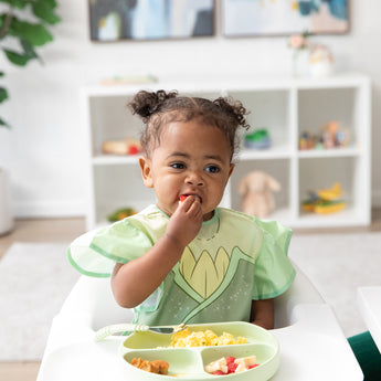 A toddler in a Bumkins Ruffle Apron Bib: Tiana eats a berry in a high chair, looking thoughtful, with toys on shelves behind.
