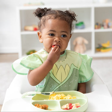 A toddler in a Bumkins Ruffle Apron Bib: Tiana eats a berry in a high chair, looking thoughtful, with toys on shelves behind.