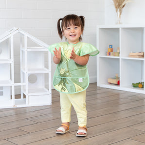 A young girl stands in a bright playroom, clapping her hands while wearing a Bumkins Ruffle Apron Bib: Tiana.