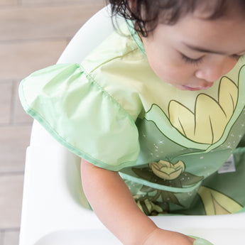 Close-up of a girl in a high chair wearing a bib from Bumkins Snack In Character 4-Piece Set: Disney Princess Magic Tiana.