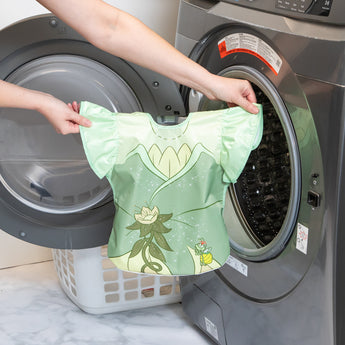 A person holds a Bumkins Ruffle Apron Bib: Tiana in front of an open front-loading washing machine.