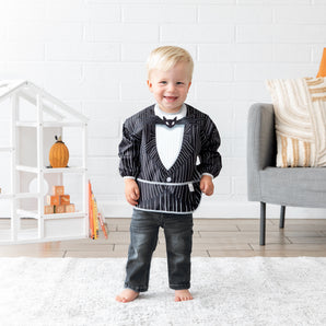 A boy wearing Bumkins Sleeved Bib: Jack Skellington stands in a bright playroom in front of a dollhouse, books, and gray chair.