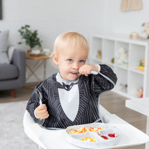 A boy wearing a bib from Bumkins Snack In Character 3-Piece Set: Disney Everyday Is Halloween Jack eats fruit from a gray plate.