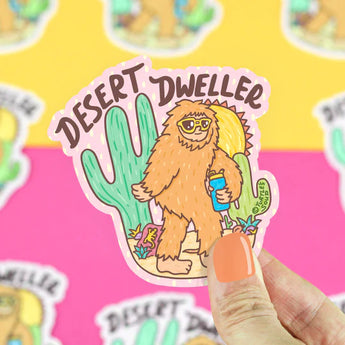 Orange-nailed hand holds Turtle Soups Desert Dweller Big Foot vinyl sticker: a scratch-resistant sasquatch with cactus and bottle.