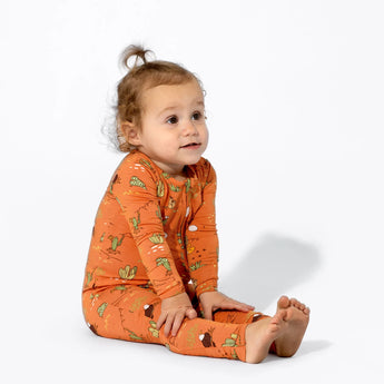 A toddler with light brown hair sits in a Bellabu Bear Bamboo Footie, Desert, gazing to the side against a plain white background.