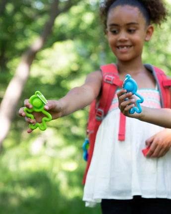 A child outdoors, wearing a red backpack, holds up green & blue toy figures with arms extended and a Fat Brain Toy Co Lil Dimpl Keychain.