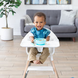 A boy beams in a high chair, wearing a Bumkins SuperBib® 3 Pack: Hangry, Dinosaurs, Blue Tropic with a blue bowl on the tray.