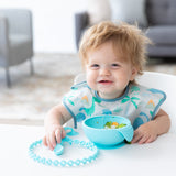 A boy with messy hair smiles, wearing a Bumkins SuperBib® 3 Pack: Hangry, Dinosaurs, Blue Tropic; a blue bowl sits on a white table in front.