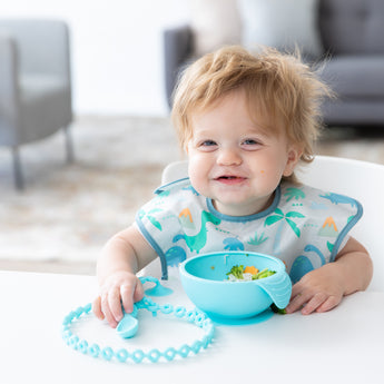 A boy with messy hair smiles, wearing a Bumkins SuperBib® 3 Pack: Hangry, Dinosaurs, Blue Tropic; a blue bowl sits on a white table in front.