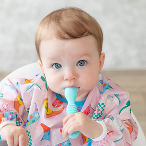 A baby girl with bright eyes in a pink patterned bib holds a Bumkins Silicone Dipping Spoons 3 Pack: Gumdrop in her mouth.