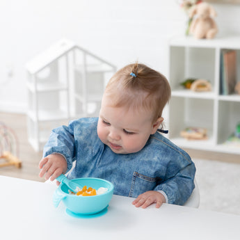 A toddler in a Bumkins Sleeved Bib: Vintage Denim eats from a blue bowl with a matching spoon at a white table in a bright playroom.