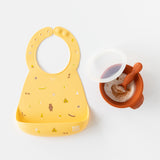 Bumkins Silicone Bib: Camp Gear lays next to a dark orange bowl of yogurt with a matching spoon & clear lid on a white background.