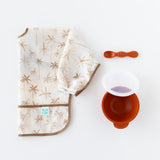 Bumkins Silicone First Feeding Set: Clay rests next to a folded beige & brown palm tree print bib on a white background.