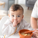 A baby sucks their thumb as an adult hand feeds them from a Bumkins Silicone First Feeding Set: Clay.