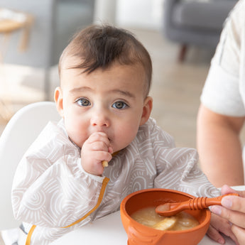 A baby sucks their thumb as an adult hand feeds them from a Bumkins Silicone First Feeding Set: Clay.