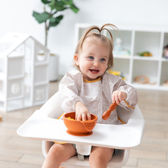 A girl in a beige bib sitting in a high chair smiles as she eats from a Bumkins Silicone First Feeding Set: Clay.