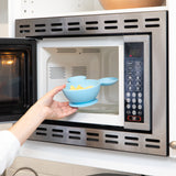 A hand places a Bumkins Silicone First Feeding Set: Mickey Mouse Light Blue filled with food into an open microwave.