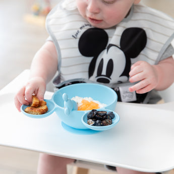 A toddler uses the Bumkins Silicone First Feeding Set: Mickey Mouse Light Blue to eat snacks on their highchair.
