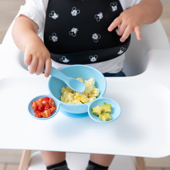 A toddler in a high chair eats scrambled eggs, tomatoes, and avocado from a Bumkins Silicone First Feeding Set: Mickey Mouse Light Blue.