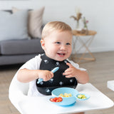 A toddler in a high chair smiles, eating from Bumkins Silicone First Feeding Set: Mickey Mouse Light Blue with chopped tomatoes and avocado.