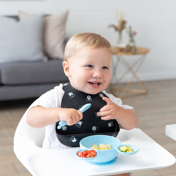 A toddler in a high chair smiles, eating from Bumkins Silicone First Feeding Set: Mickey Mouse Light Blue with chopped tomatoes and avocado.
