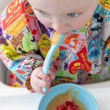 A baby in a colorful bib enjoys applesauce with a spoon from Bumkins Silicone First Feeding Set: Mickey Mouse Light Blue.