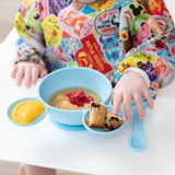 A child wearing a colorful bib enjoys applesauce, fruit, and a muffin from a Bumkins Silicone First Feeding Set: Mickey Mouse Light Blue.