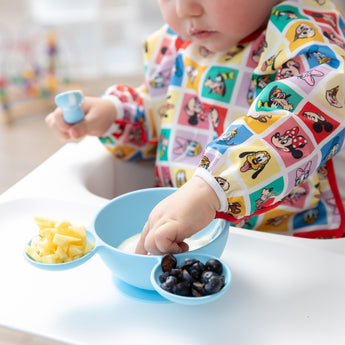 Close-up of a child in a colorful bib exploring self-feeding with Bumkins Silicone First Feeding Set: Mickey Mouse Light Blue.