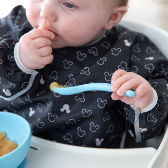 A baby in high chair wearing a patterned bib eats from Bumkins Silicone First Feeding Set: Mickey Mouse Light Blue.