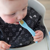 A baby in a high chair wears a bib, holding a spoon from Bumkins Silicone First Feeding Set: Mickey Mouse Light Blue.
