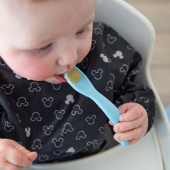 A baby in a high chair wears a bib, holding a spoon from Bumkins Silicone First Feeding Set: Mickey Mouse Light Blue.