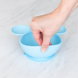 A hand lifts a Bumkins Silicone First Feeding Set: Mickey Mouse Light Blue on a marble surface, showcasing the suction cup bottom.