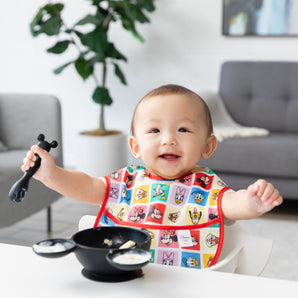 Bumkins Silicone First Feeding Set: Mickey Mouse Classic Black sits on a table in front of a baby in a colorful bib in a cozy living room.