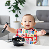 Bumkins Silicone First Feeding Set: Mickey Mouse Classic Black sits on a table in front of a baby in a colorful bib in a cozy living room.