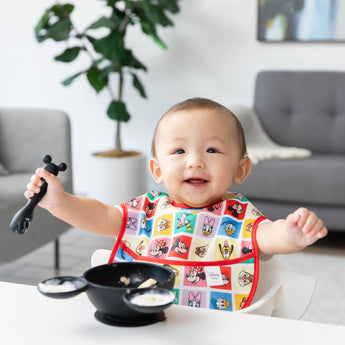 Bumkins Silicone First Feeding Set: Mickey Mouse Classic Black sits on a table in front of a baby in a colorful bib in a cozy living room.