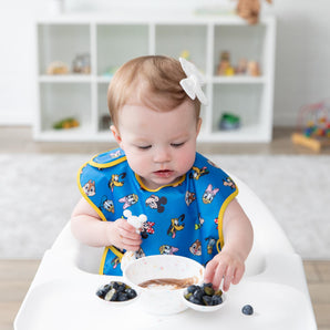 A girl wearing a blue Disney-themed bib in a high chair enjoys berries from Bumkins Silicone First Feeding Set: Mickey Mouse Vanilla Sprinkle 