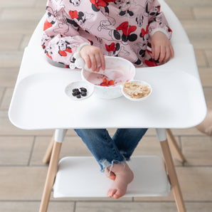 A child in a bib enjoys yogurt with berries from a Bumkins Silicone First Feeding Set: Mickey Mouse Vanilla Sprinkle on a high chair tray.