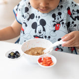 A child in a Mickey Mouse bib eats oatmeal with a spoon from a Bumkins Silicone First Feeding Set: Mickey Mouse Vanilla Sprinkle at a table.