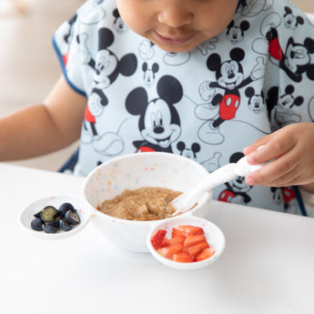 A child in a Mickey Mouse bib eats oatmeal with a spoon from a Bumkins Silicone First Feeding Set: Mickey Mouse Vanilla Sprinkle at a table.