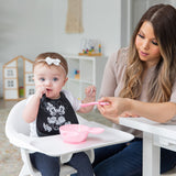 A woman feeds a baby in a high chair with a Bumkins Silicone Dipping Spoons: Minnie Mouse (Black and Pink) from a pink bowl on the tray.
