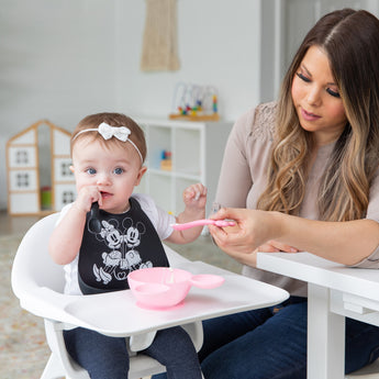 A woman feeds a baby in a high chair with a Bumkins Silicone Dipping Spoons: Minnie Mouse (Black and Pink) from a pink bowl on the tray.