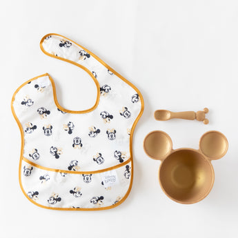 Bumkins Silicone First Feeding Set: Minnie Mouse Gold rests next to a black & white Minnie Mouse-themed bib on a white background.