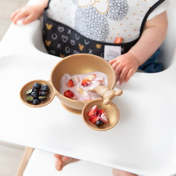Close-up of Bumkins Silicone First Feeding Set: Minnie Mouse Gold with yogurt and berries in front of a child in a black & white bib.